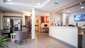 A hotel lobby with a reception desk, vending machines, a seating area, and a television screen. - Ace hôtel Annemasse Genève (Ville-la-Grand)