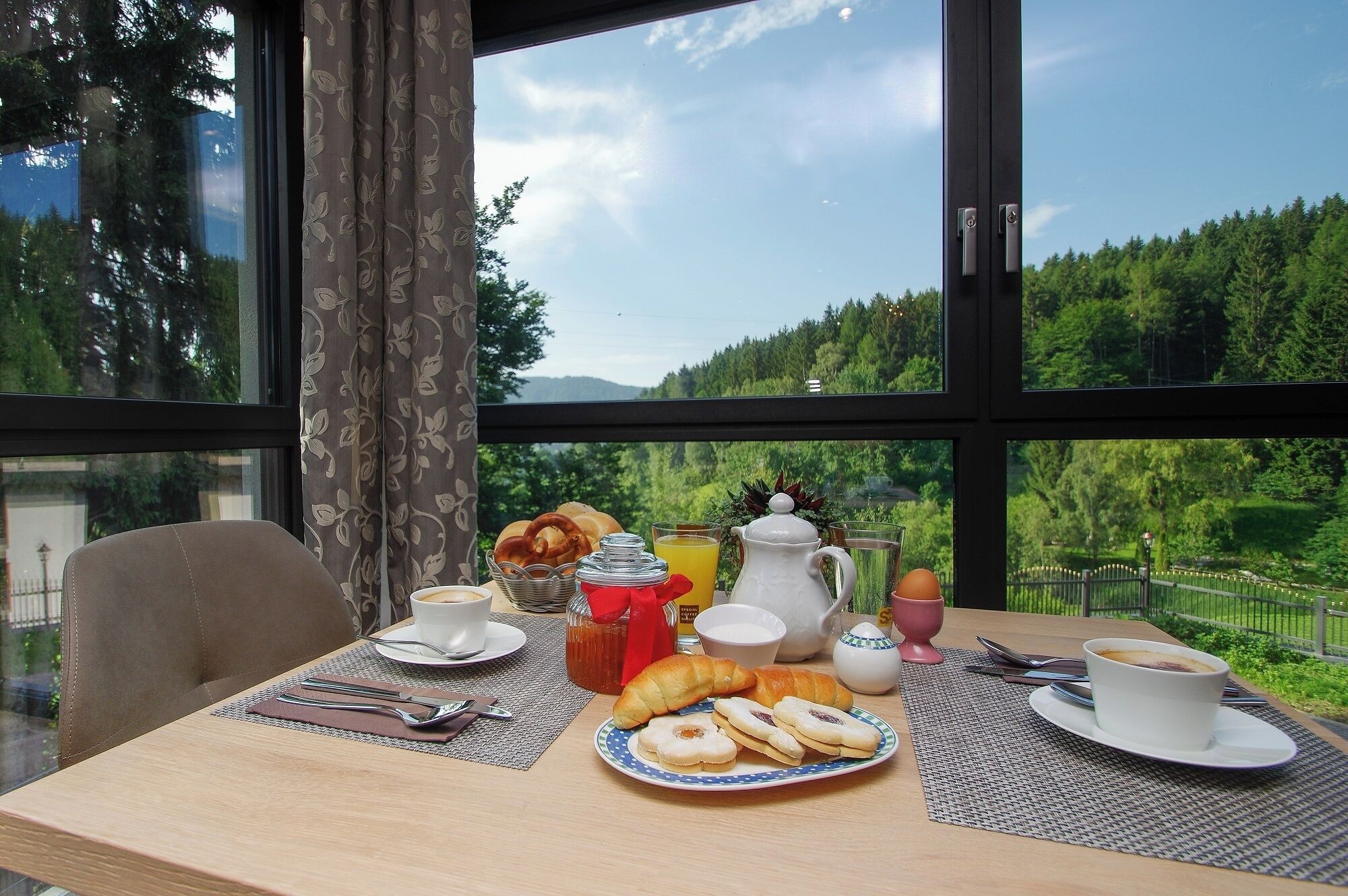 Una mesa de desayuno con pasteles, café y jugo, con vistas a árboles y montañas.