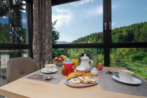 Una mesa de desayuno con pasteles, café y jugo, con vistas a árboles y montañas.