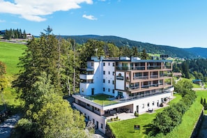 Un hotel de varios pisos con terraza en techo plano, rodeado de vegetación y colinas.