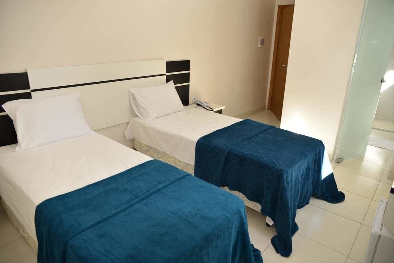 A hotel room with two single beds, each with a white mattress and a blue bedspread. There is a bedside table with a phone on it, and a door leading to another room.