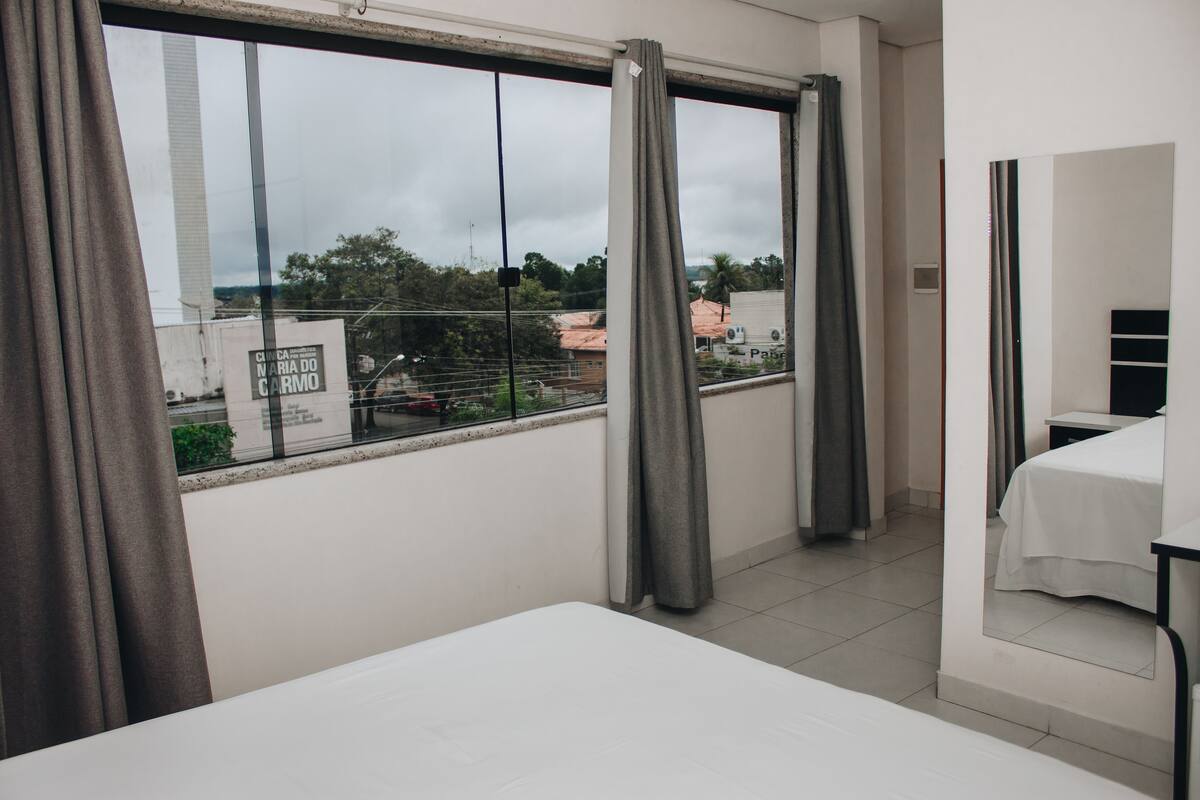 A hotel room with a large window, a bed, a small table, and a mirror.
