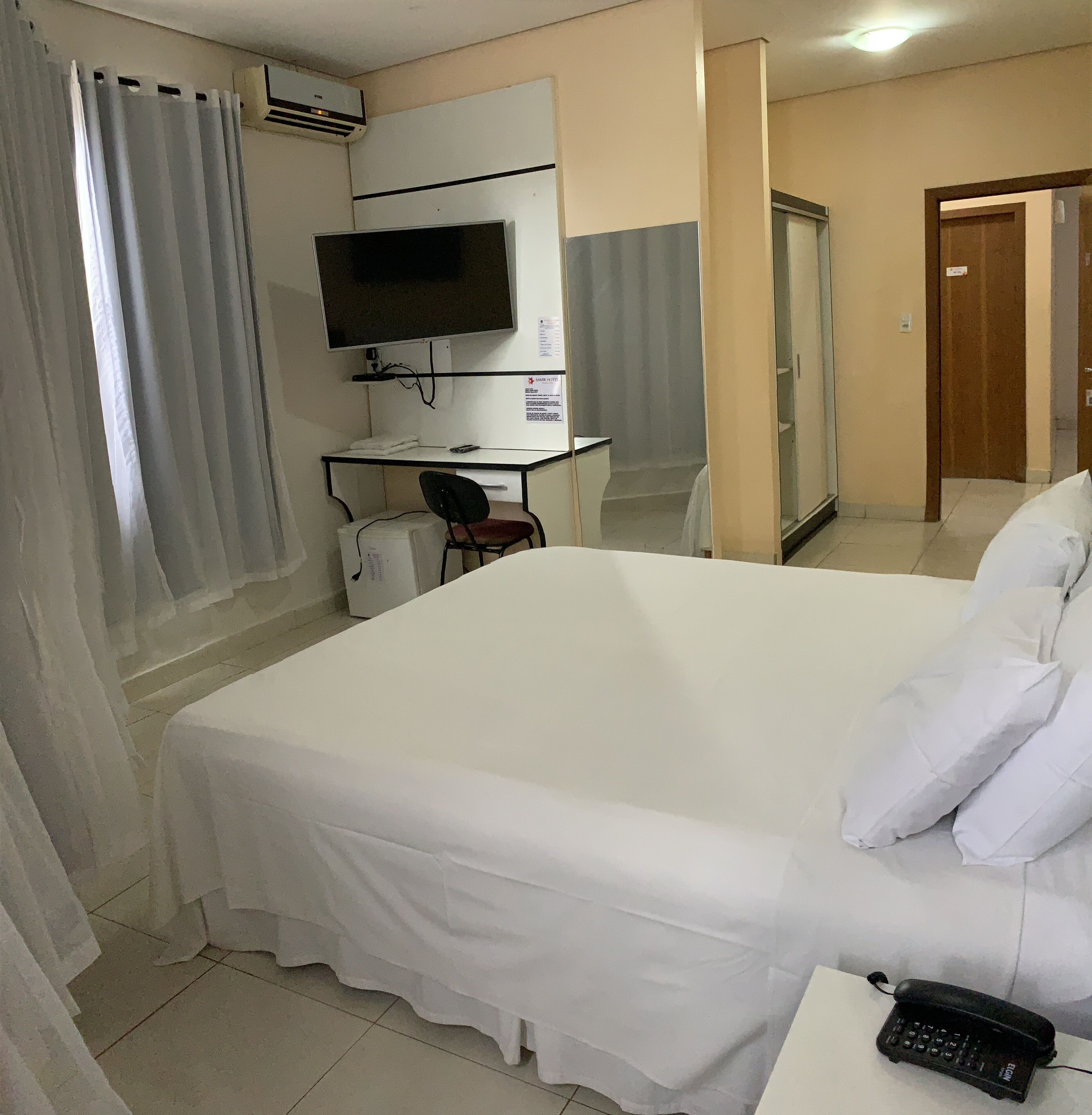Ett hotellrum med en säng, ett skrivbord med en dator, en stol, en telefon och en luftkonditioneringsanläggning.