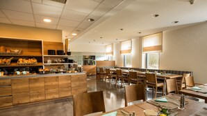 A modern dining area with wooden tables and chairs, a central counter with bread and pastries, and large windows with blinds. - Hotel der Freiraum (Guessing)