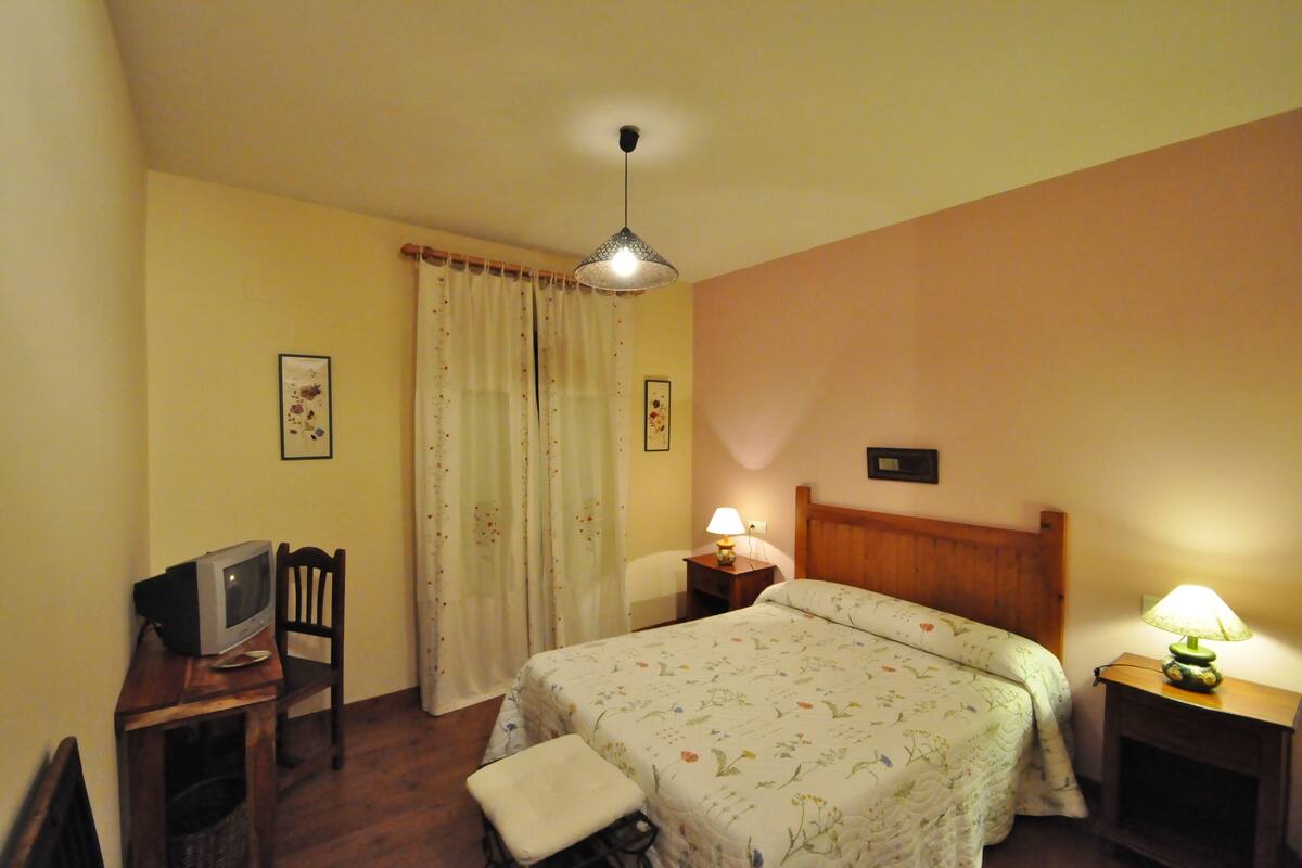 A bedroom with a bed, a chair, a small table with a lamp, a television on a stand, and a window with curtains.