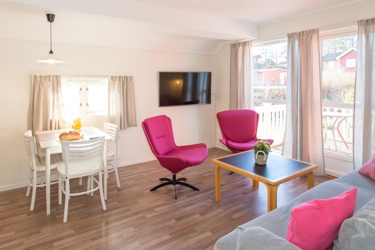 A modern living room with a dining area, a flat-screen TV, and a balcony with a view of houses.