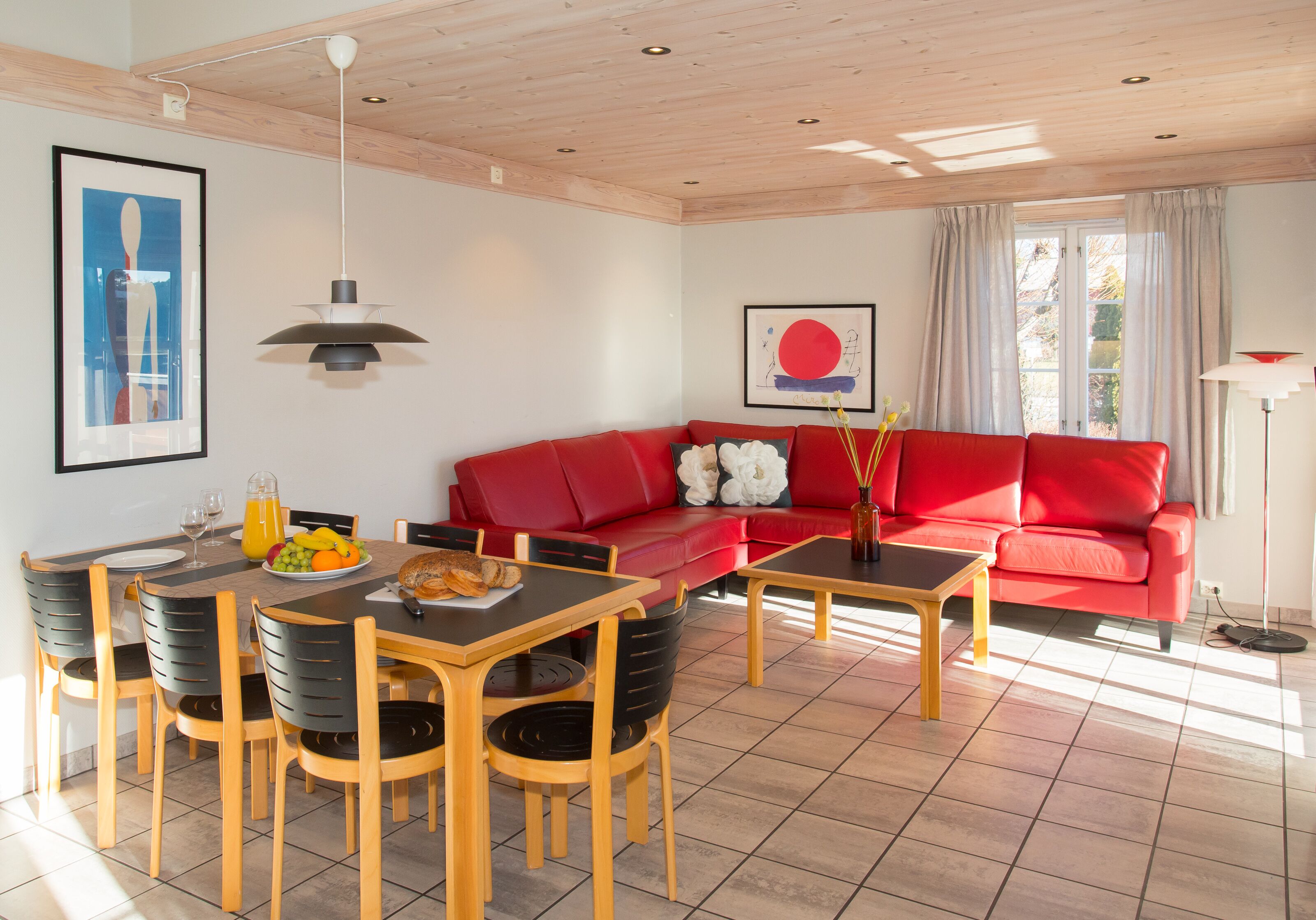 Una sala de estar moderna con un sofá rojo, una mesa de comedor de madera y sillas. Se observa una mesa de comedor con una fuente de frutas y una botella de jugo de naranja.