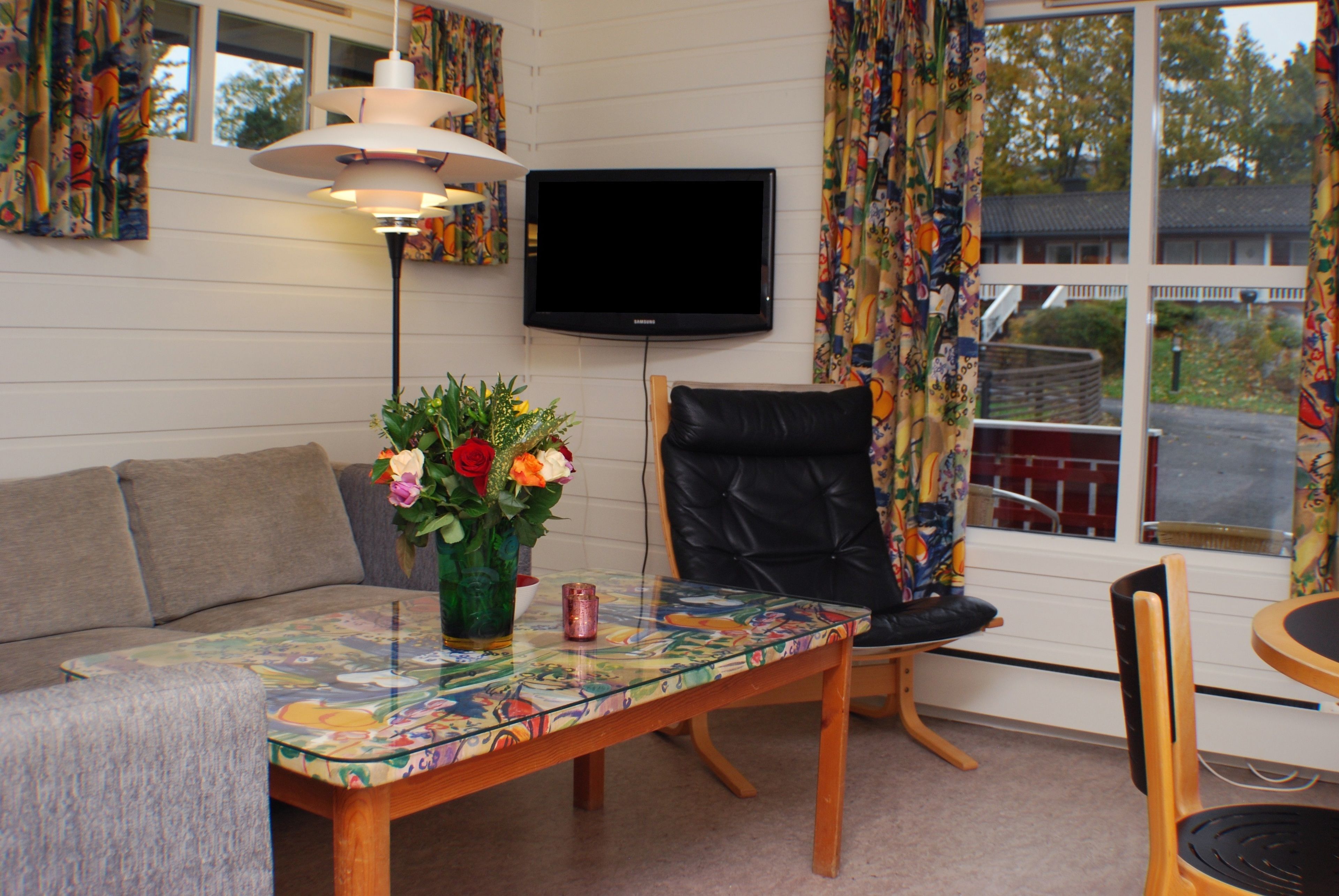 A living room with a sofa, a table with a floral tablecloth, a black leather chair, a TV mounted on the wall, and a window with curtains.