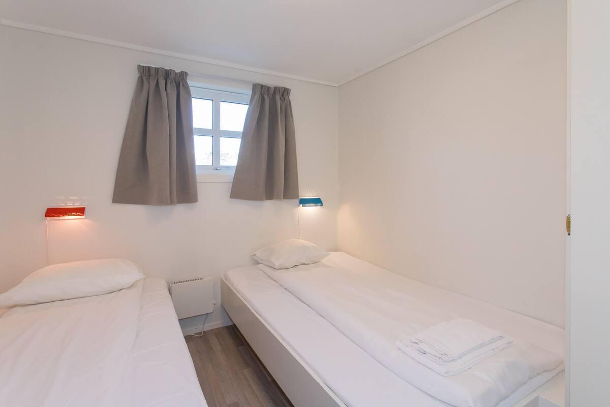 A small, clean room with two single beds, a window with curtains, and wall-mounted reading lights.