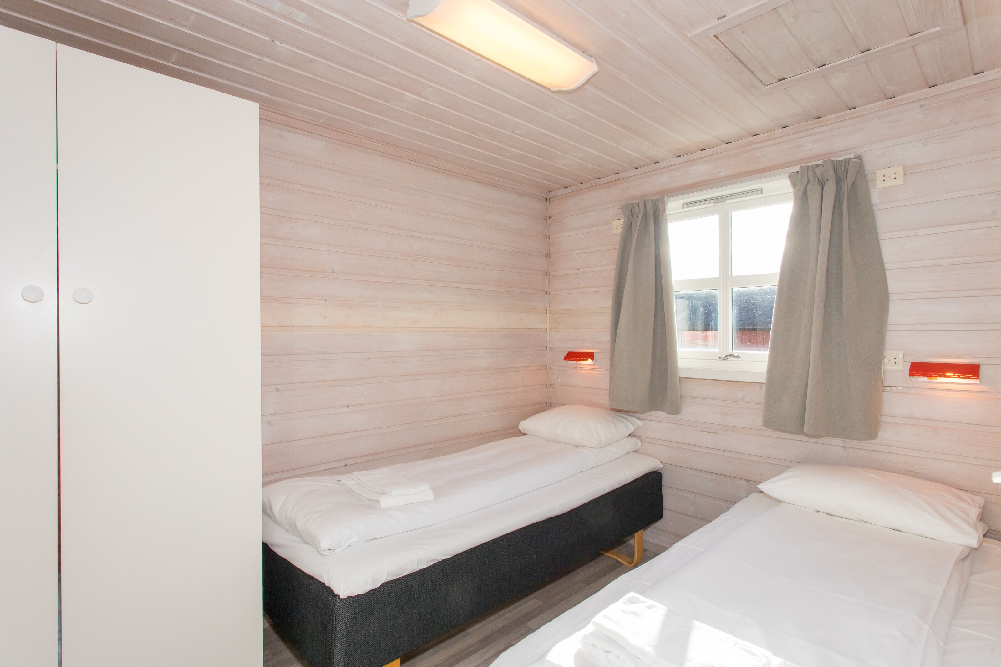 A small, single-room hotel room with two beds, wooden walls, a window with curtains, and a wall-mounted light fixture.