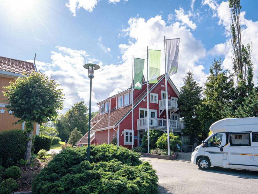 Um edifício vermelho com varanda, cercado por vegetação e uma caravana estacionada.