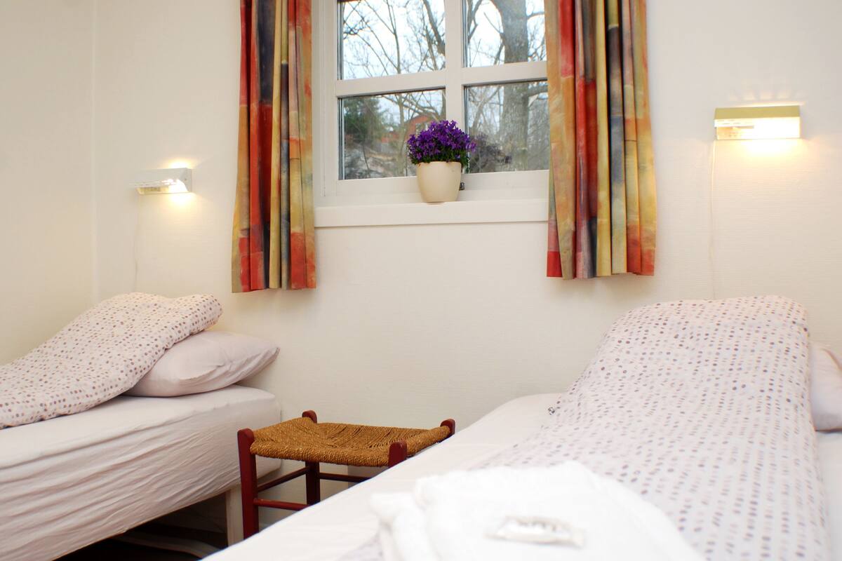 A room with two beds, a small chair, a window with curtains, and a potted plant on the windowsill.