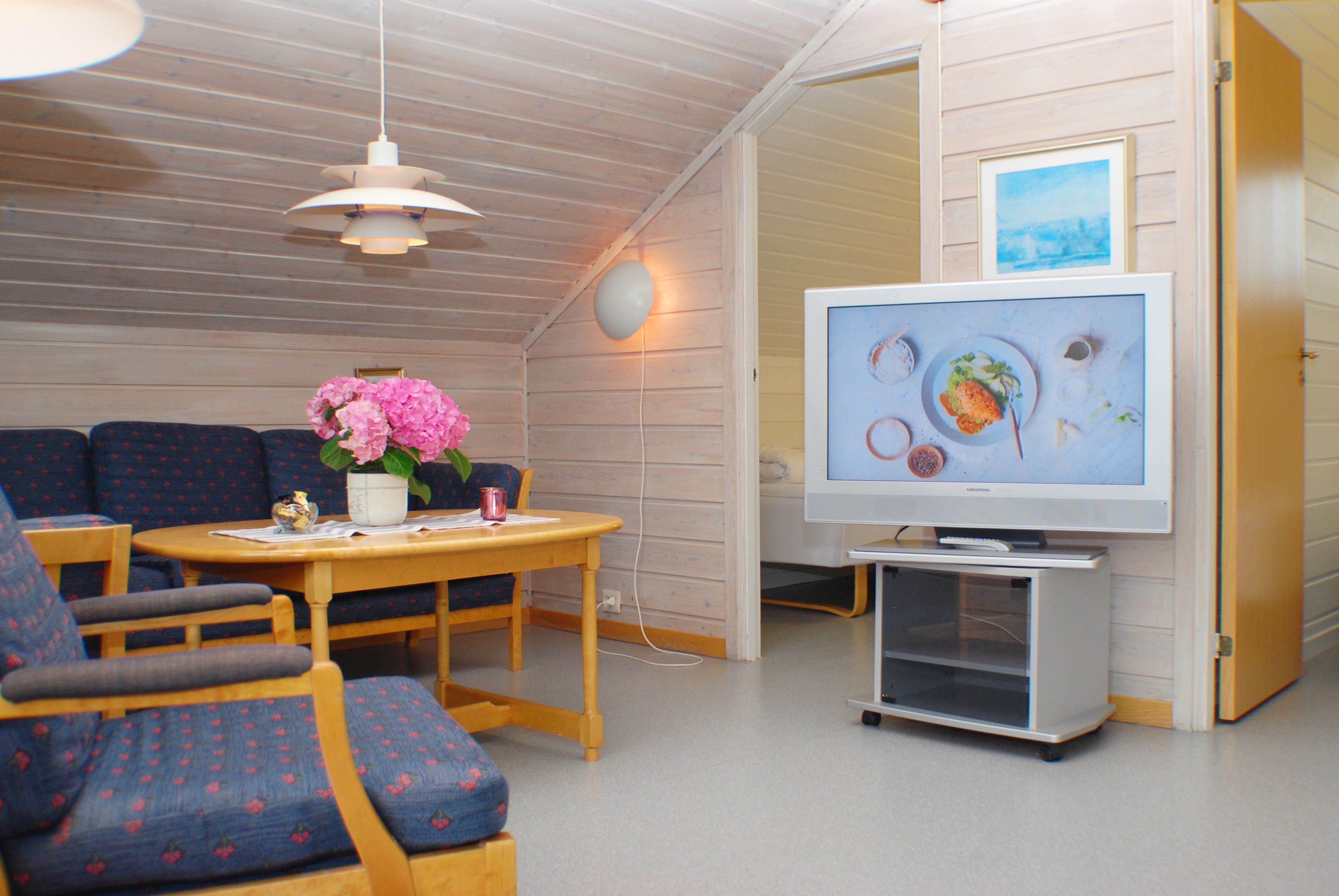 Una habitación acogedora con techo de madera, televisor sobre un mueble bajo, una mesa con una silla y un sofá.