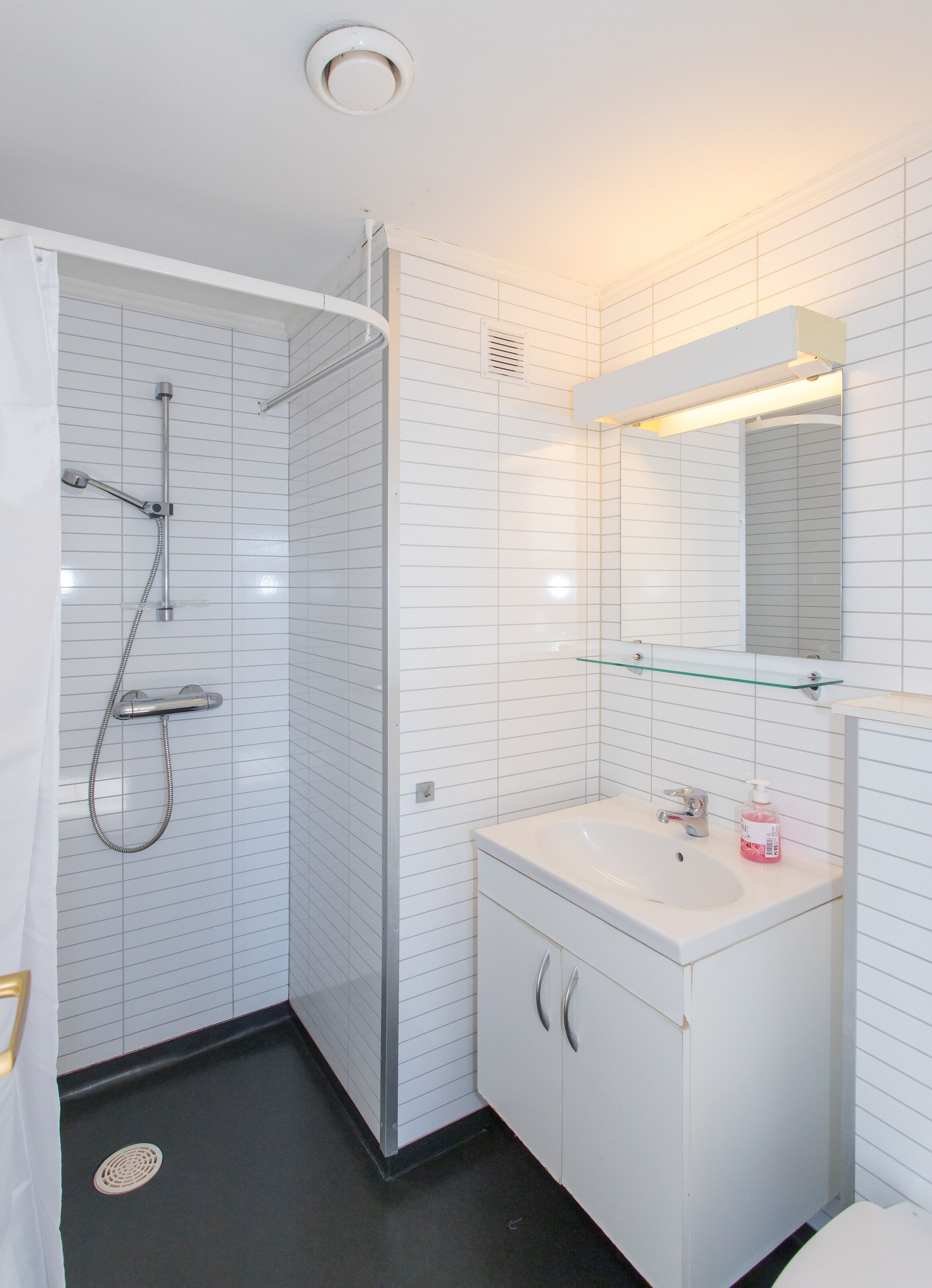 A modern bathroom with white tiled walls, a glass shower enclosure, a white sink with a mirror above it, and a black floor.