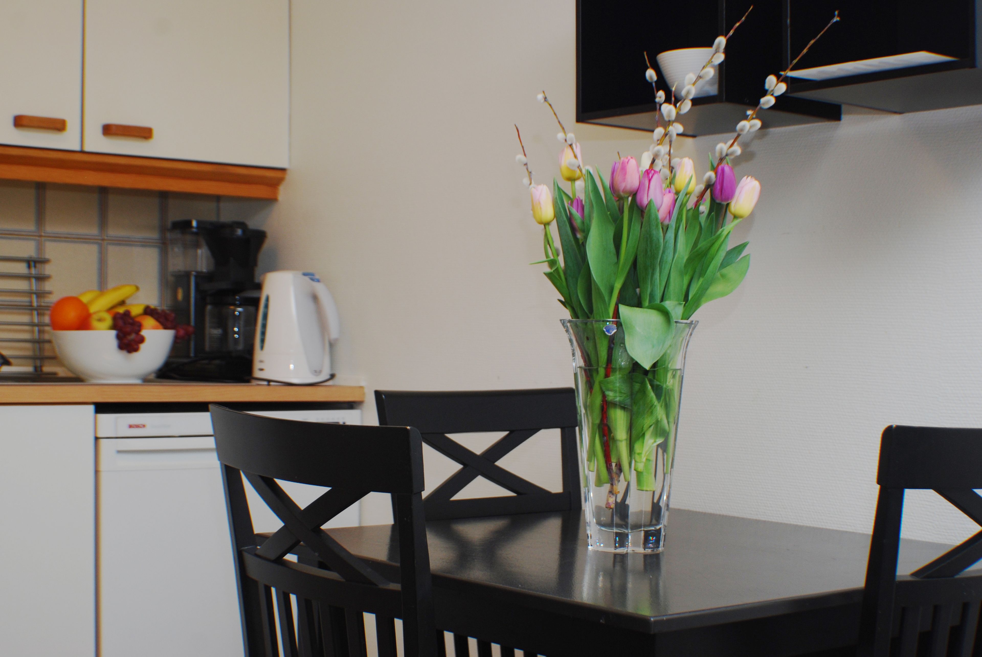 A dining table with a clear vase containing flowers and a bowl of fruit.
