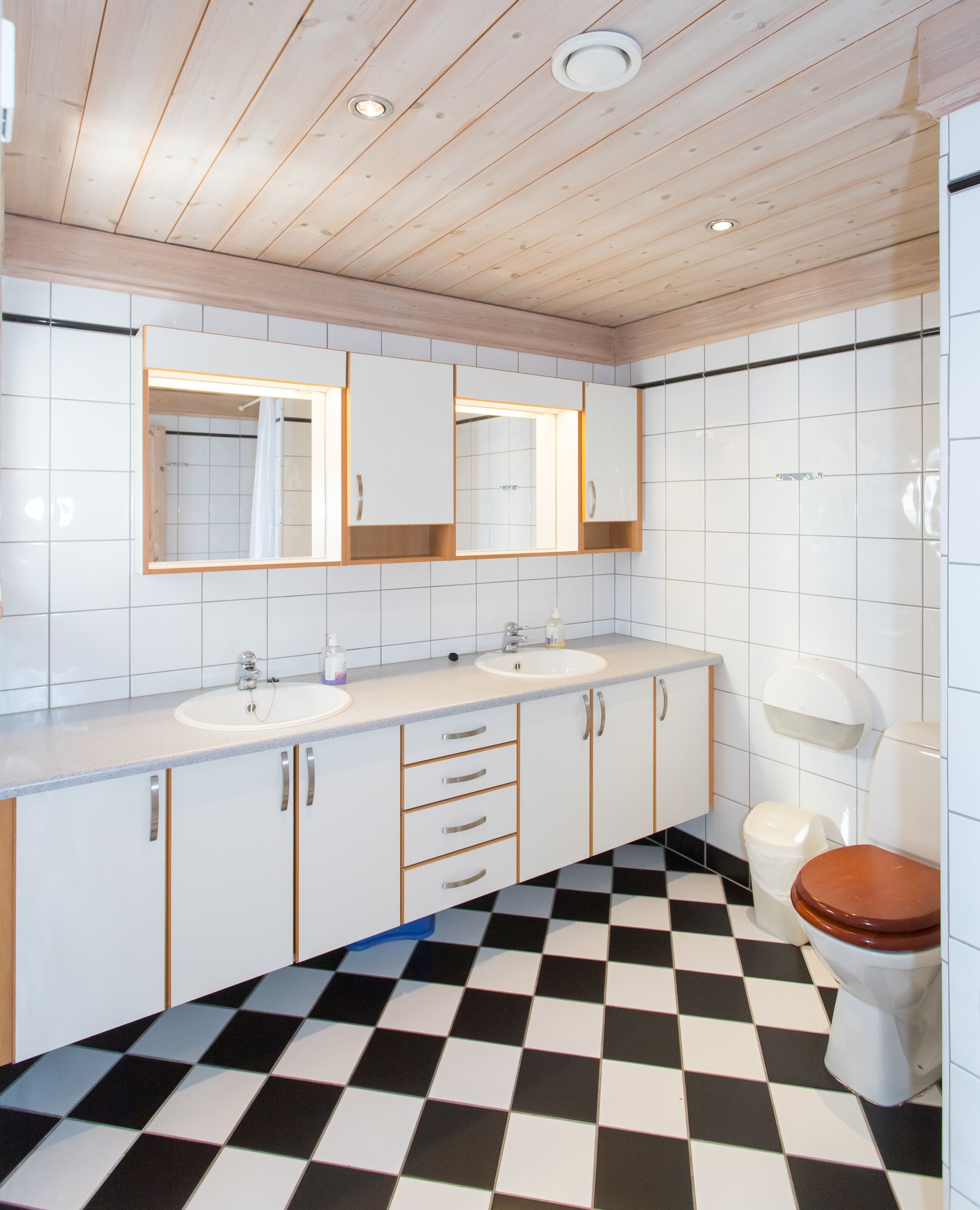 Un baño con piso a cuadros, azulejos blancos, gabinetes de madera, un lavamanos, un espejo y un inodoro.
