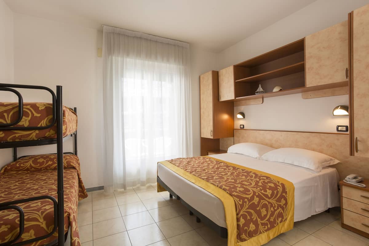 A hotel room with bunk beds, a large bed with a patterned bedspread, a wardrobe with shelves, a bedside table with a lamp, and a window with curtains.