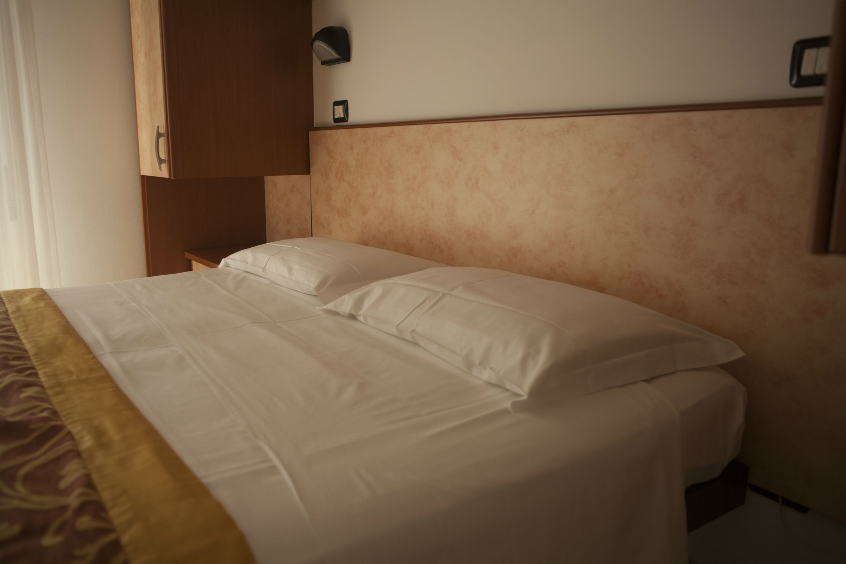 A hotel room with a bed, a wooden wardrobe, and a wall-mounted light fixture.