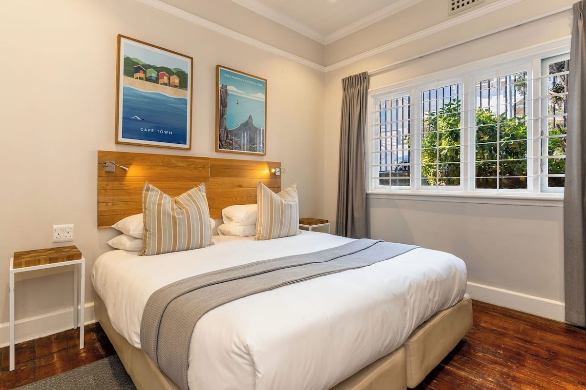 A neatly arranged bedroom with a large bed, a wooden headboard, two framed pictures on the wall, a bedside table, and a window with curtains.