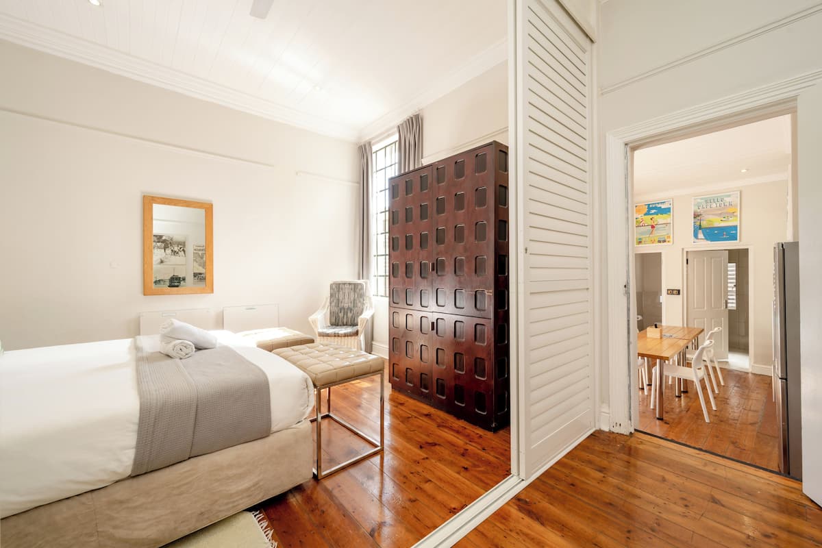 A hotel room with a bed, a chair, a wooden cabinet, and a dining area visible through an open door.