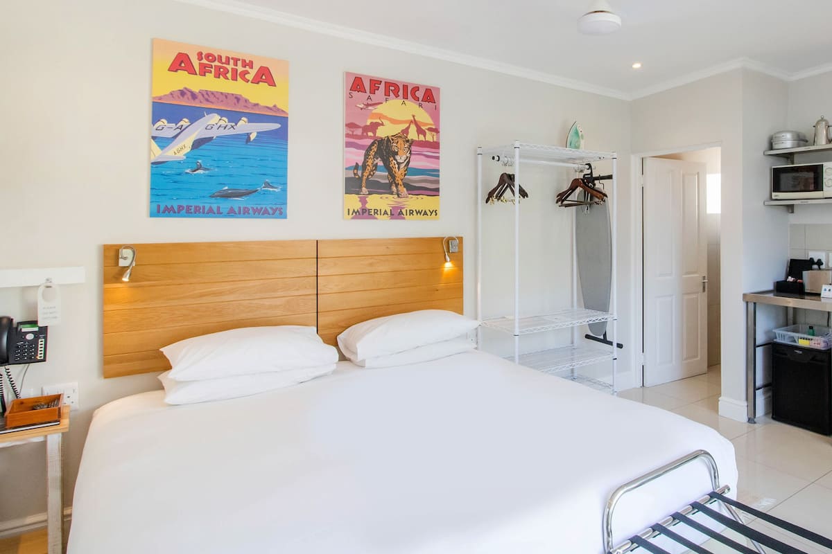 A hotel room with a large bed, a wooden headboard, a wardrobe, a kitchenette, and two posters on the wall.