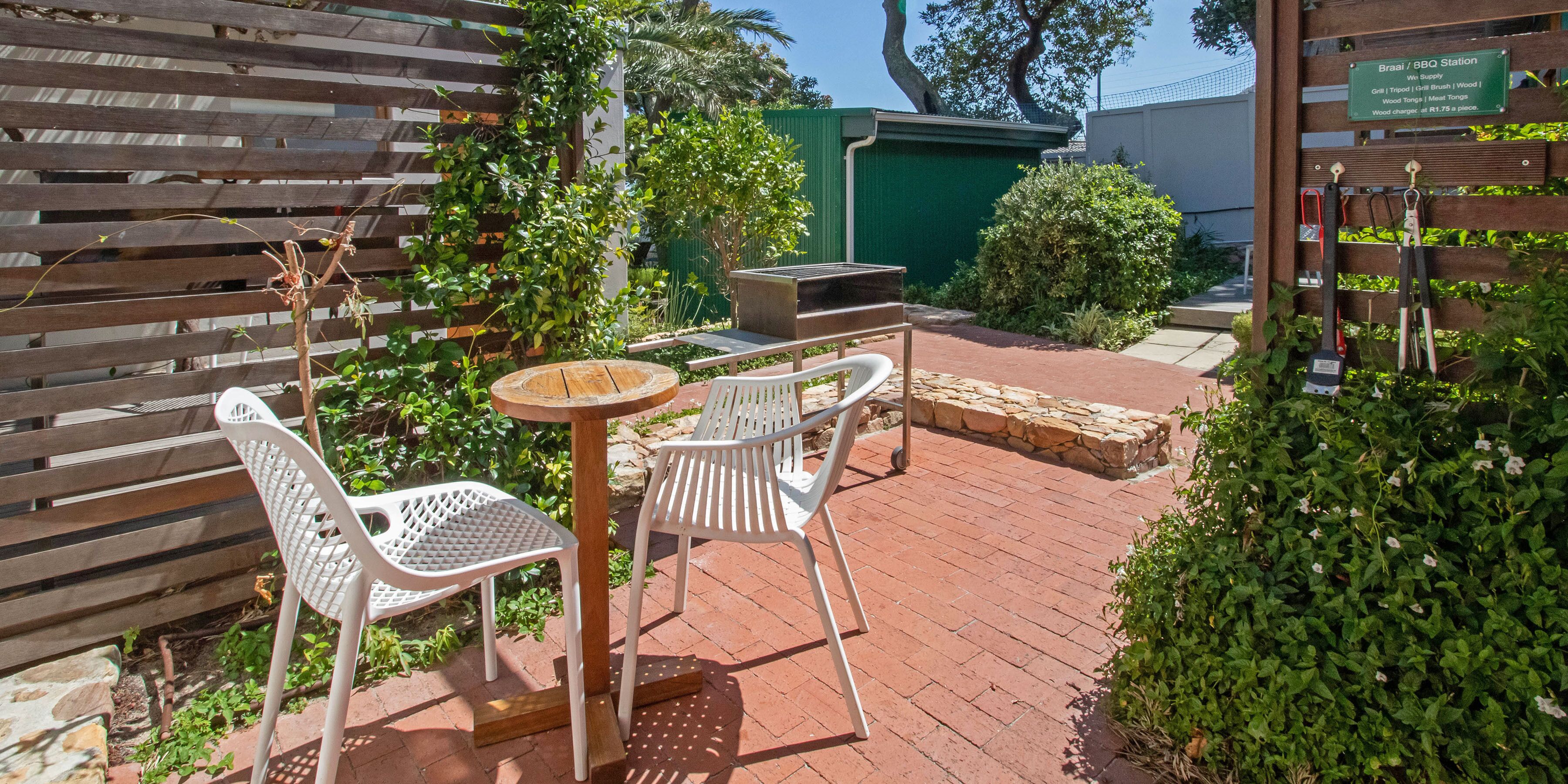 Un espace patio extérieur avec une table ronde et deux chaises blanches, entouré de verdure et d’un mur en briques.