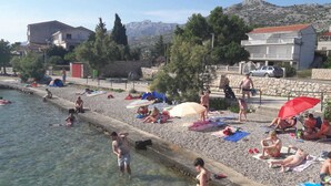 A pebbled beach with people sunbathing and swimming, surrounded by buildings and trees. - Hotel Barba (Starigrad)