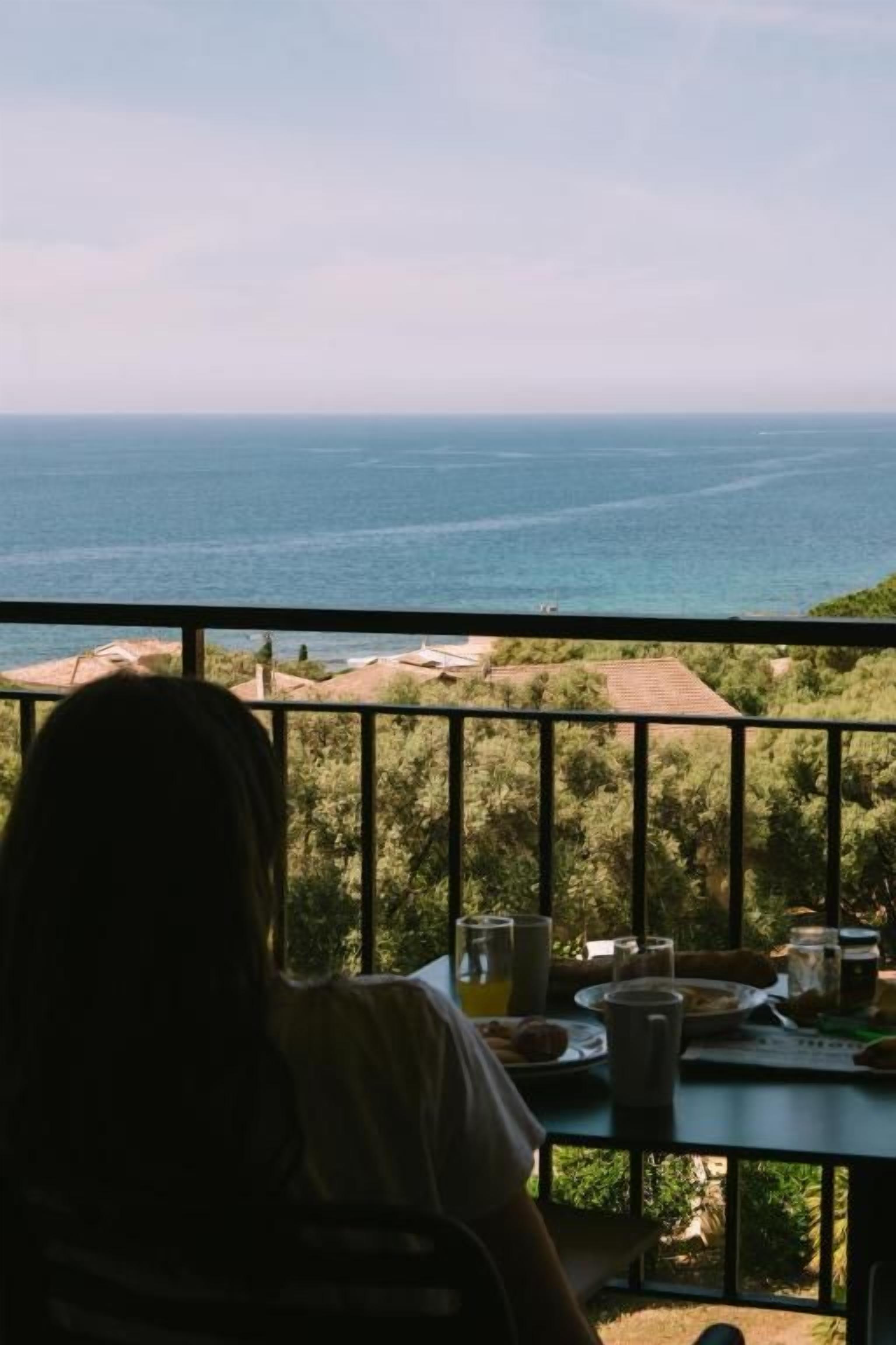 A person seated at a table with a view of the sea, a railing, and greenery in the background.