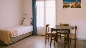 Une chambre à coucher avec un lit, une fenêtre avec des rideaux, une table à manger avec des chaises et un tableau accroché au mur.