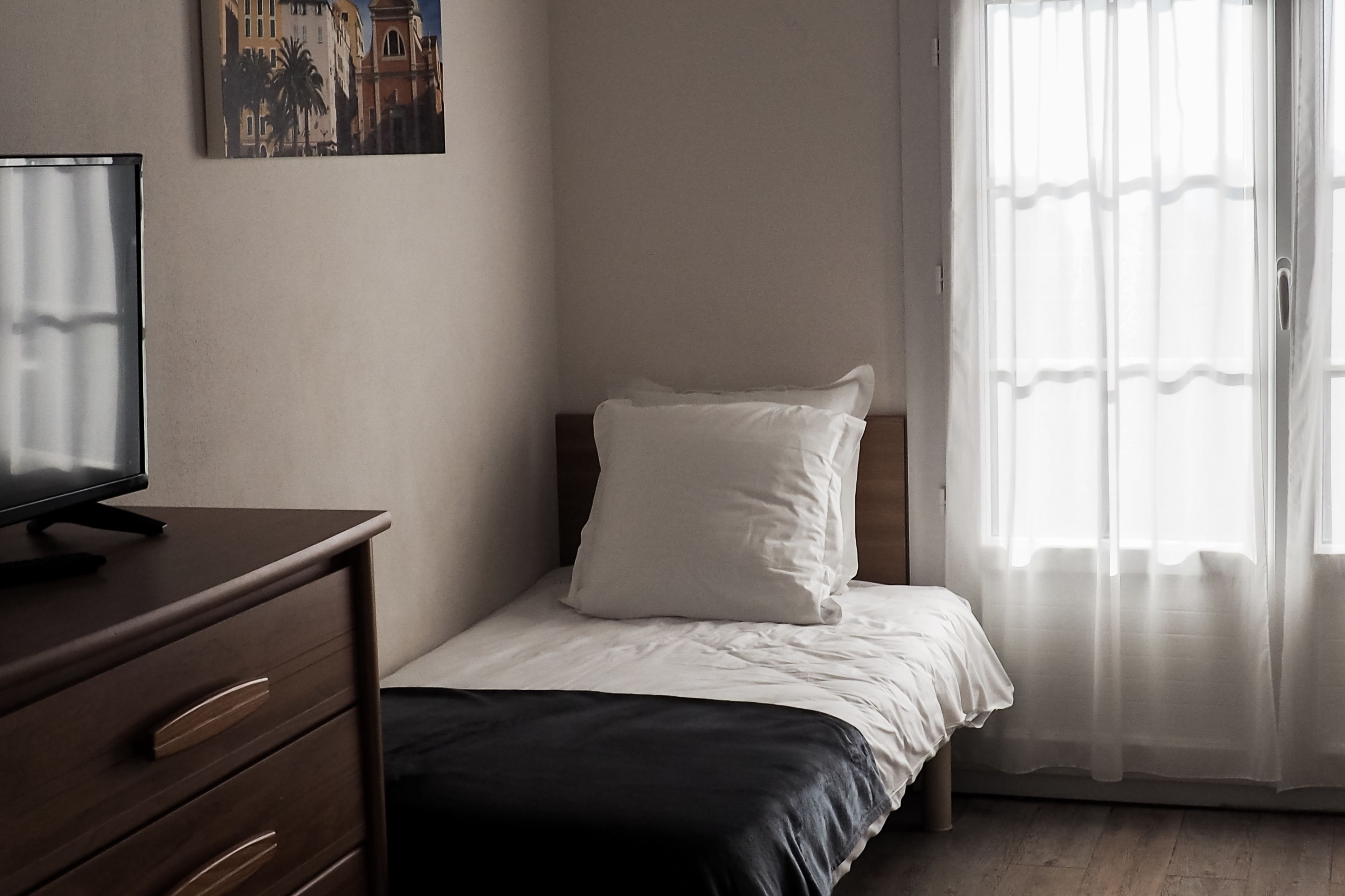 Een slaapkamer met een bed, een dressoir, een televisie op het dressoir en een raam met dunne gordijnen.