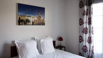 Une chambre d’hôtel avec un lit, une table de chevet avec une lampe, un tableau représentant un paysage urbain et des rideaux à motifs.