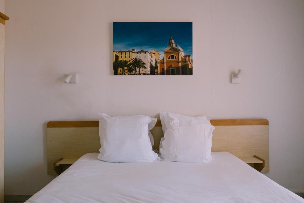 Une chambre d’hôtel avec un lit, deux oreillers blancs, une tête de lit en bois et un tableau représentant un bâtiment accroché au mur.