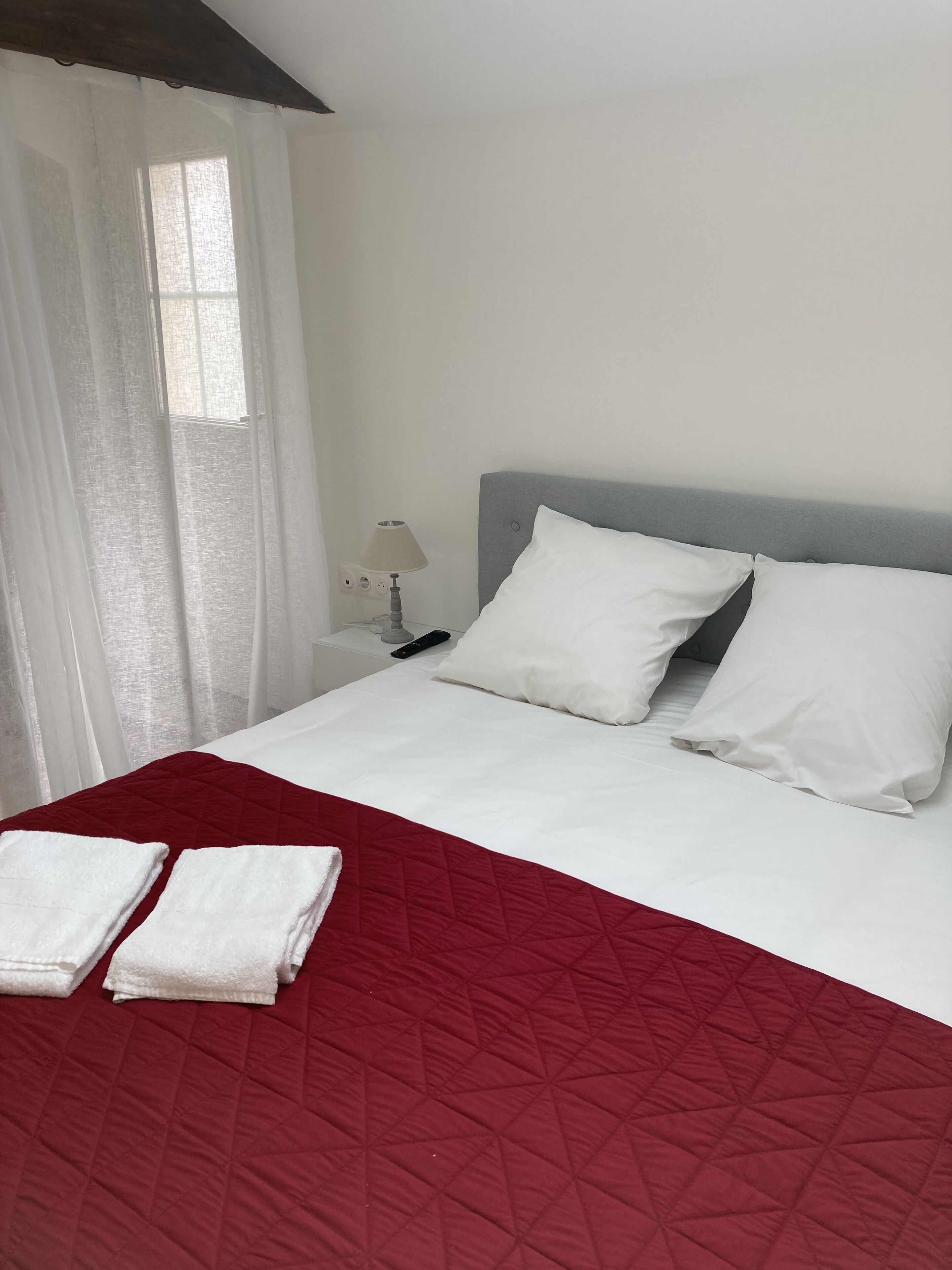 A neatly made bed with white linens and a red quilted bedspread. There are two white towels folded at the foot of the bed. A bedside table with a lamp and a remote control is visible.
