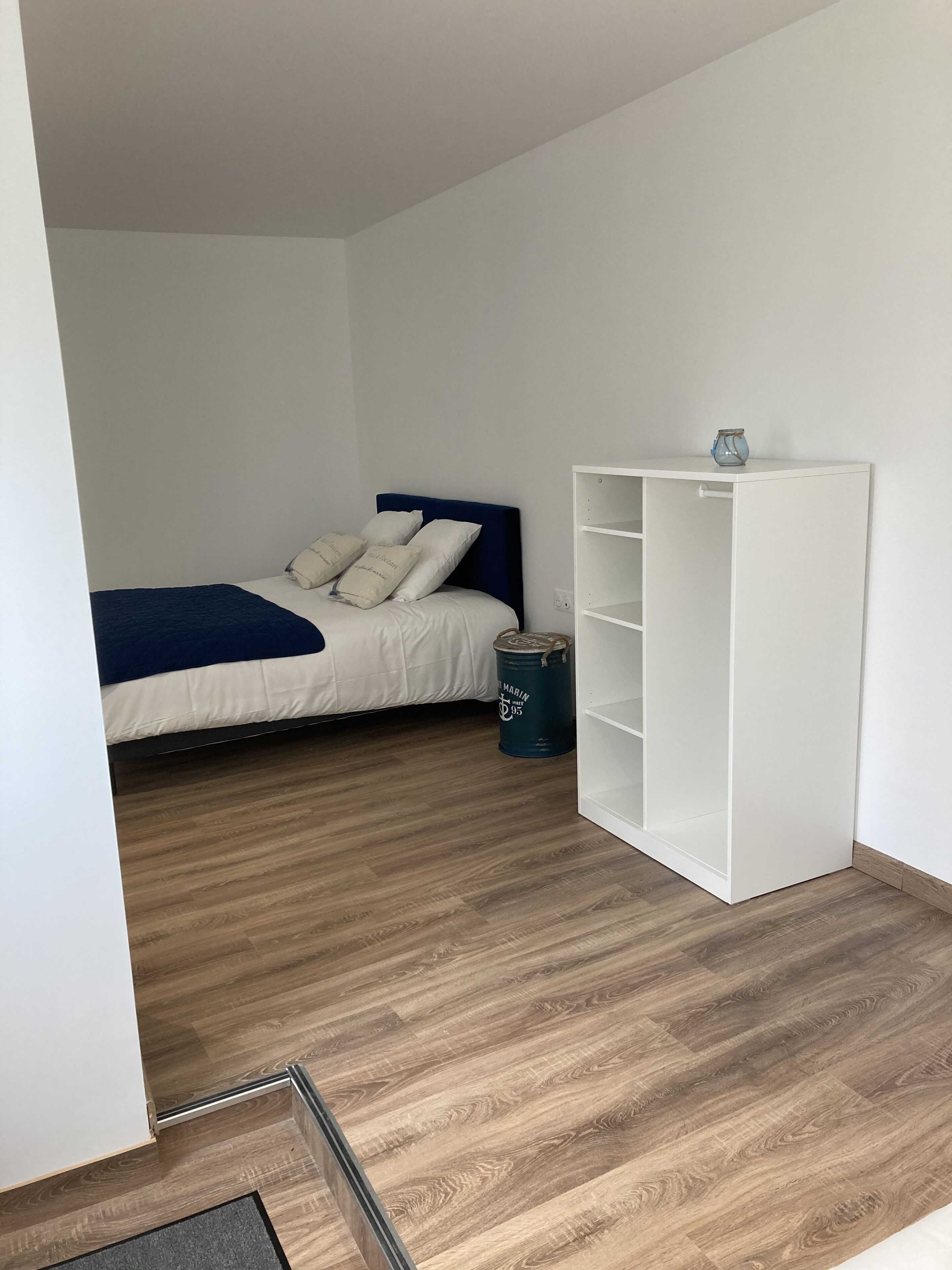 A bedroom with a bed, a white wardrobe, and a blue trash bin.