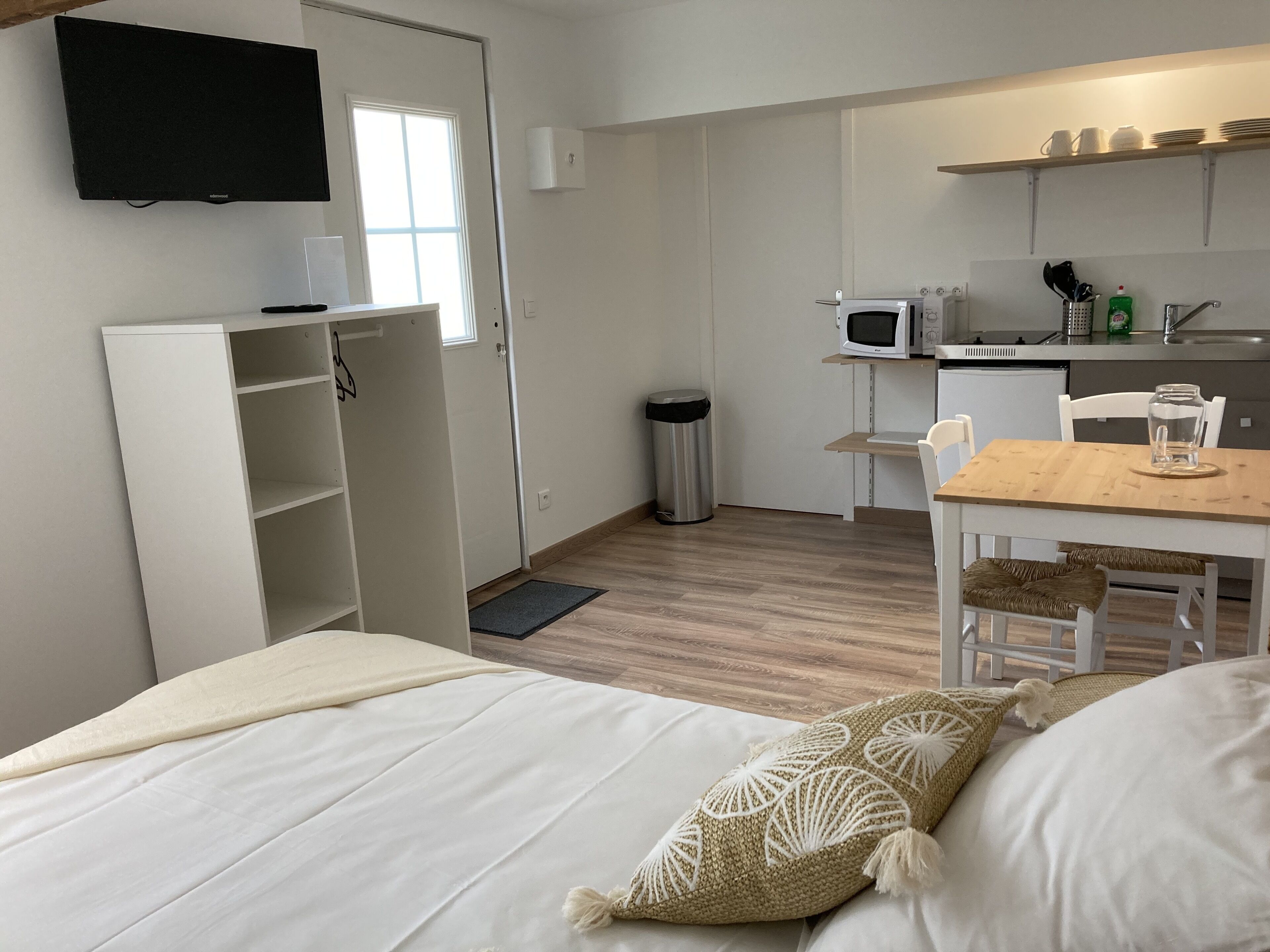 A compact room with a bed, a small kitchenette, a TV mounted on the wall, and a window.
