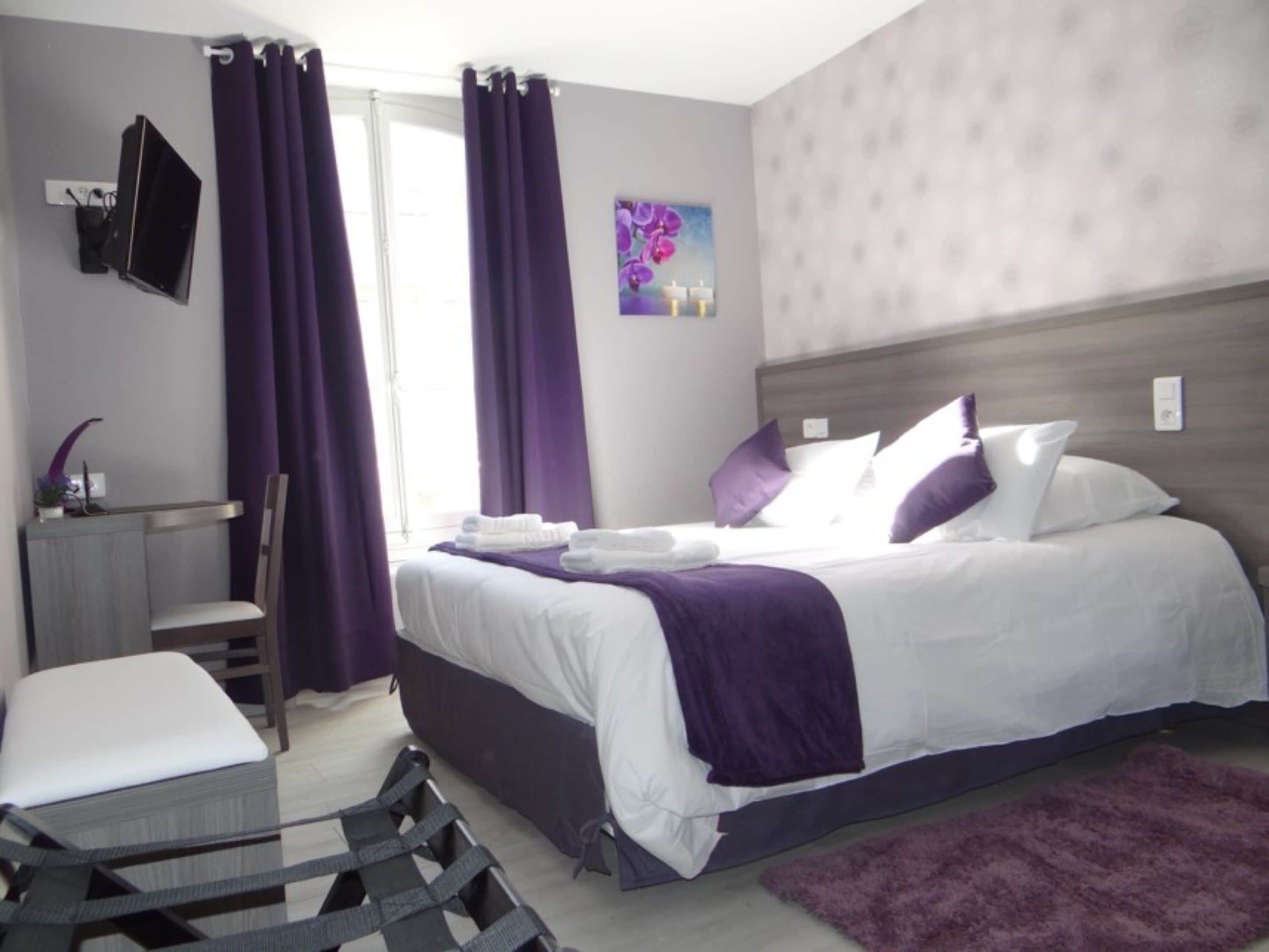 Een hotelkamer met een bed, een bureau, een stoel, een televisie en een raam met paarse gordijnen.