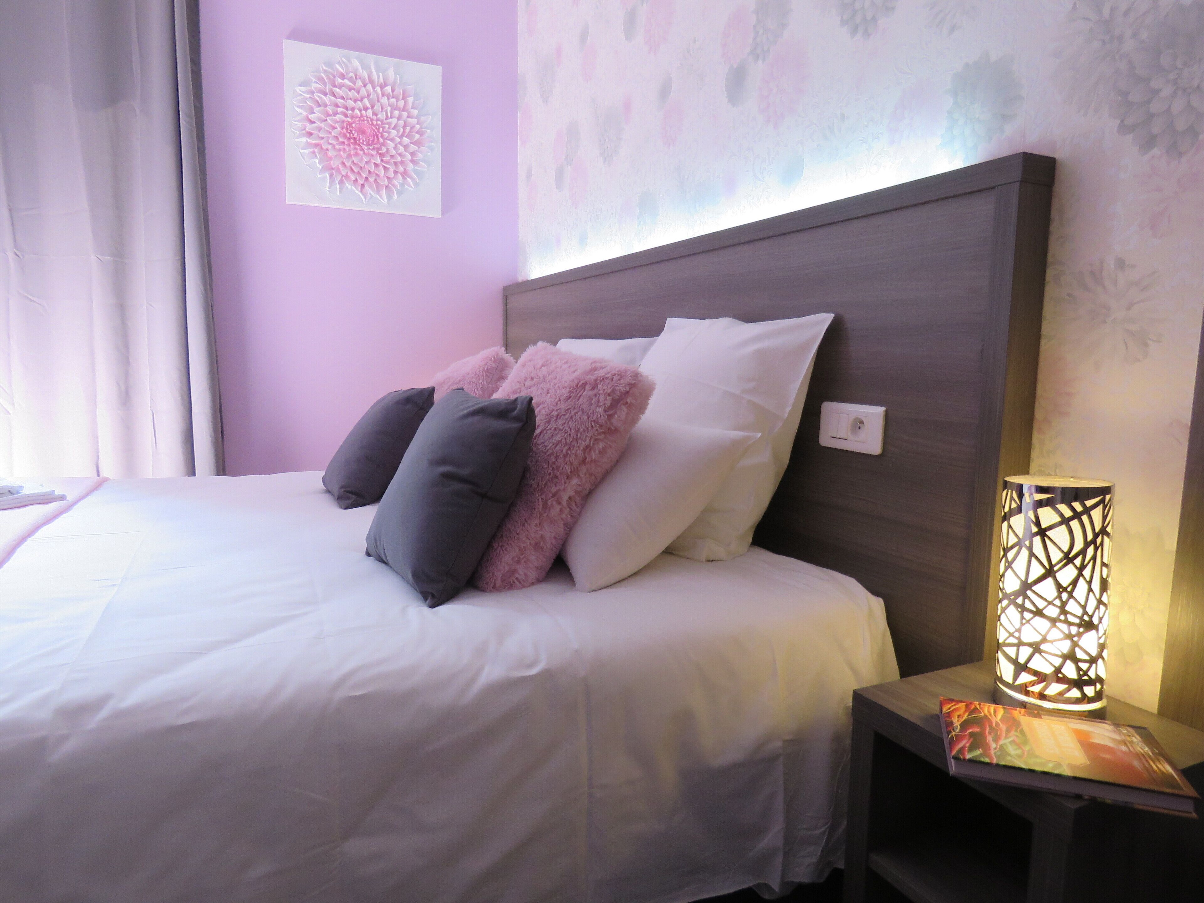 Una cama bien hecha con almohadas blancas y grises, un plaid rosa suave y una mesita de noche con una lámpara decorativa.