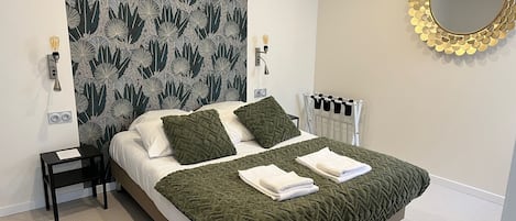 A neatly made bed with a green blanket and pillows, a bedside table with a lamp, a wall-mounted mirror, and a radiator.