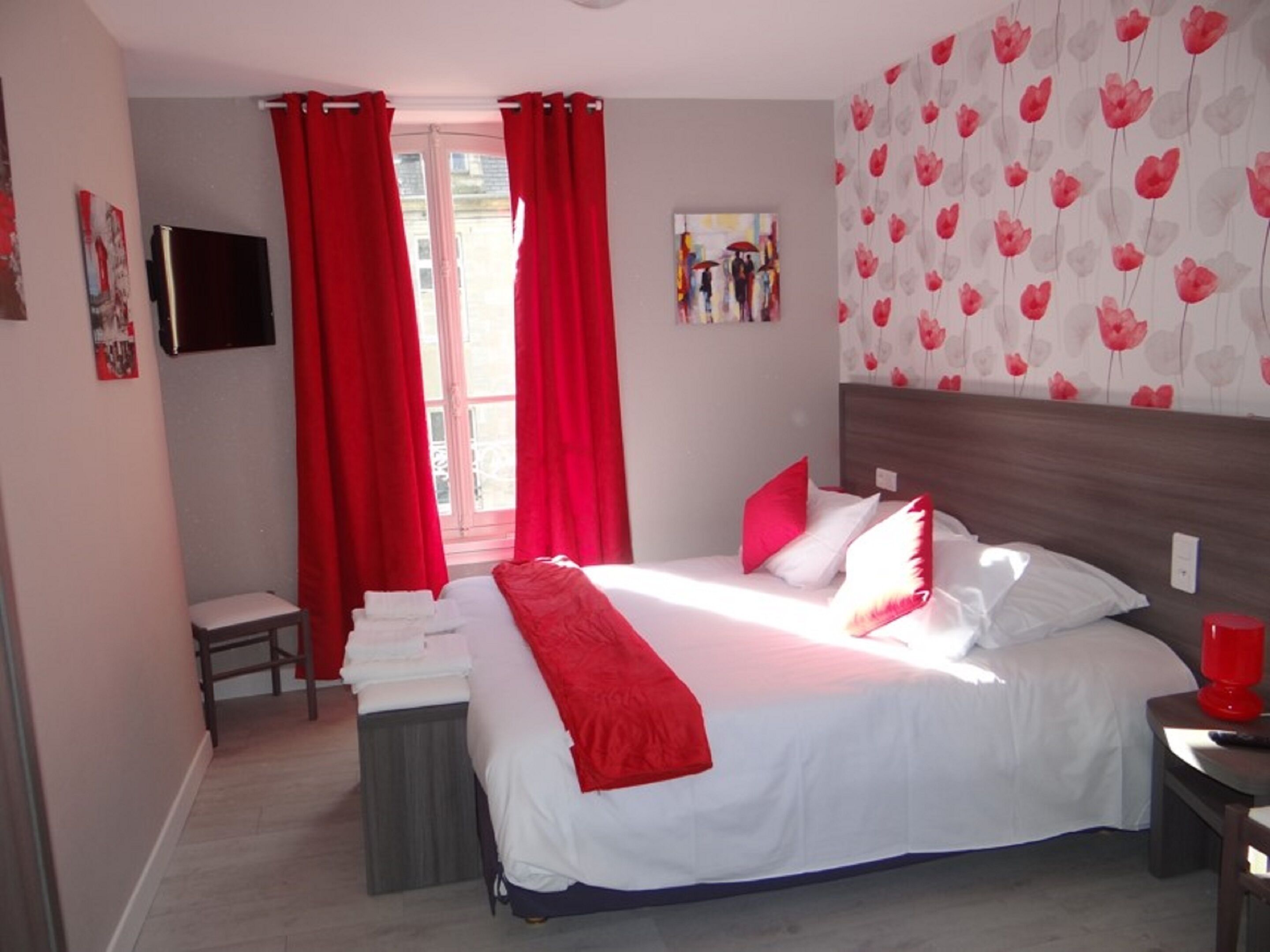 A bedroom with a bed, red curtains, a small table with towels, a wall-mounted TV, and a floral wallpaper.