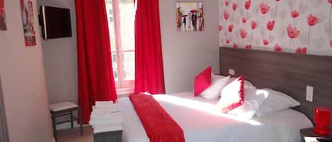 A bedroom with a bed, red curtains, a small table with towels, a wall-mounted TV, and a floral wallpaper.