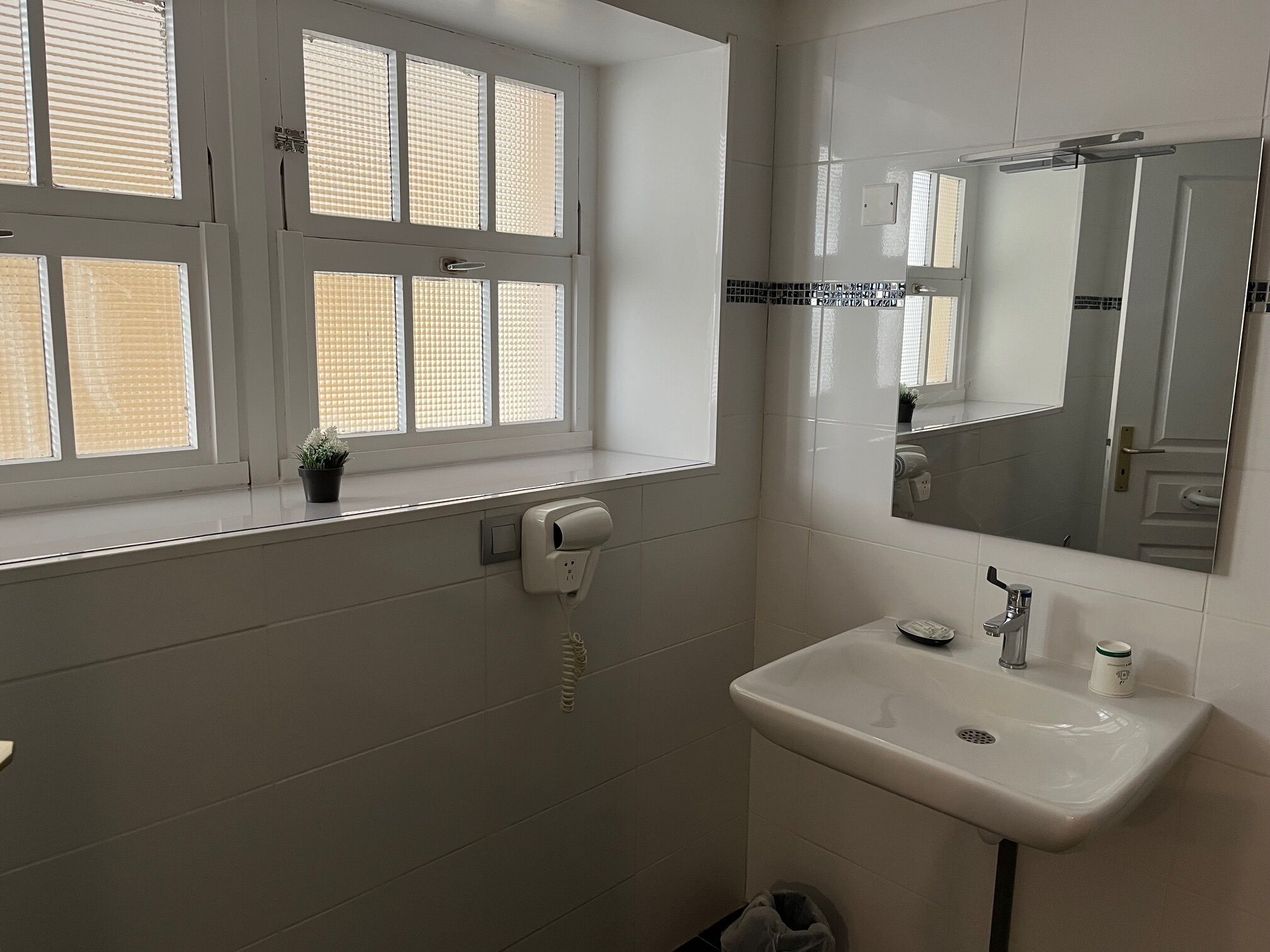 A bathroom with a window, a sink, a mirror, and a hand dryer.
