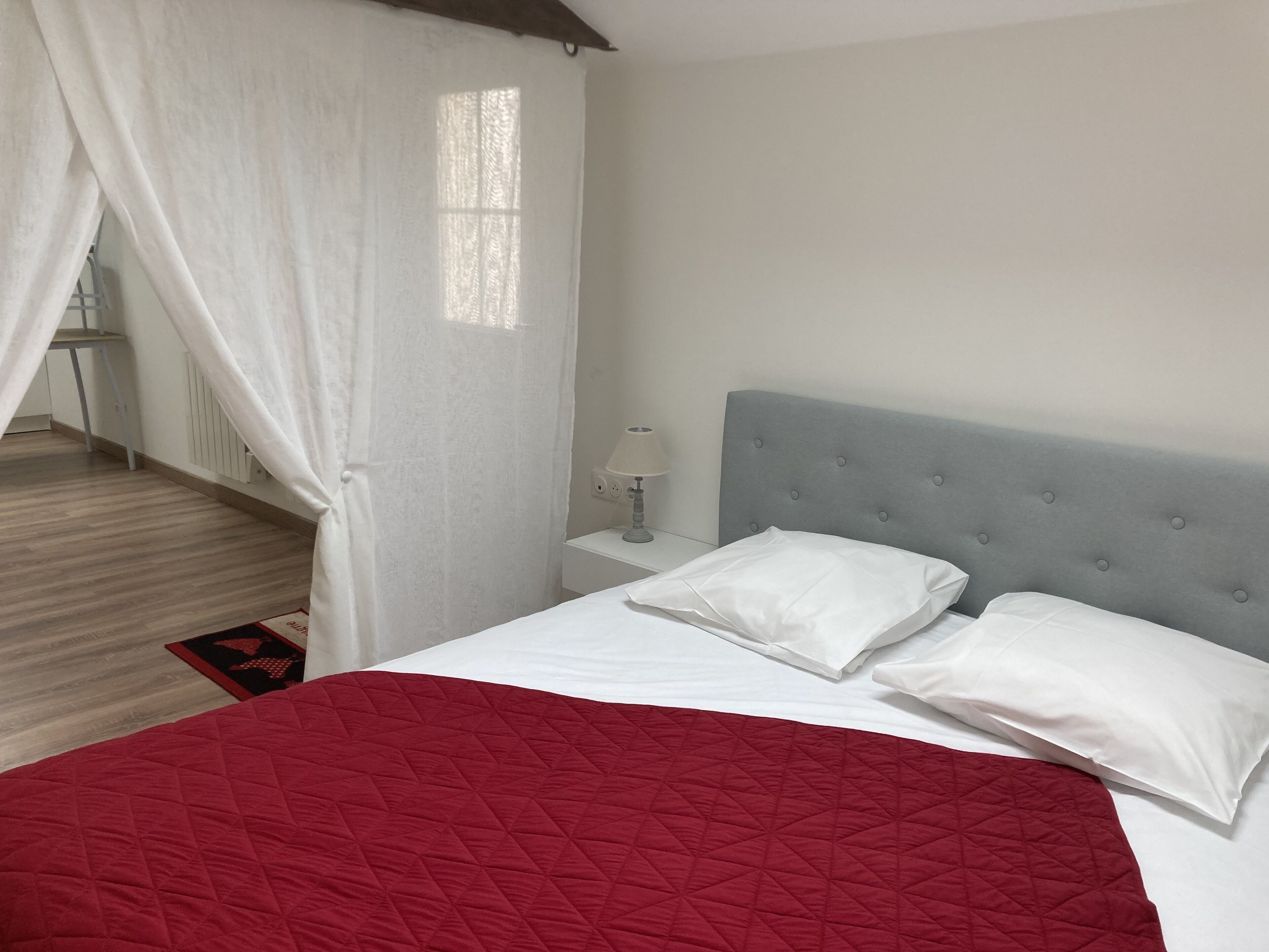 Un dormitorio con cama, cortinas blancas, una mesita de noche y una colcha acolchada roja.