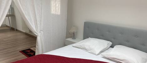 A bedroom with a bed, white curtains, a nightstand, and a red quilted bedspread.