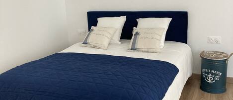 A bed with a navy blue quilt, two pillows with text, a dark blue headboard, and a small trash can with text.