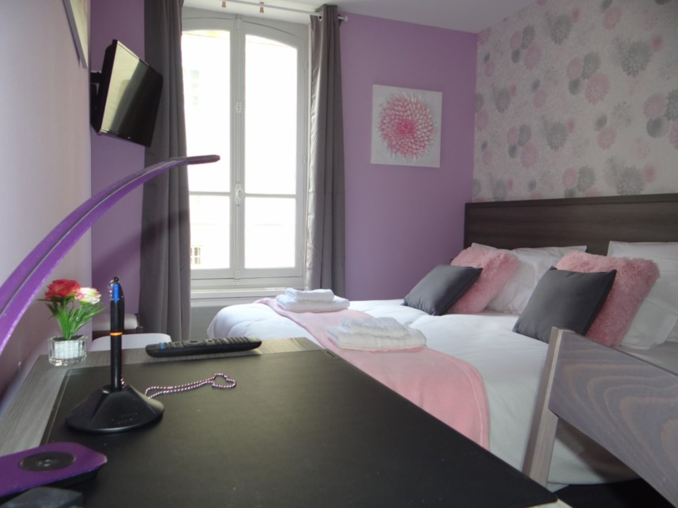 Une chambre d’hôtel avec un lit, un bureau doté d’une lampe, une télécommande et un vase avec des fleurs.