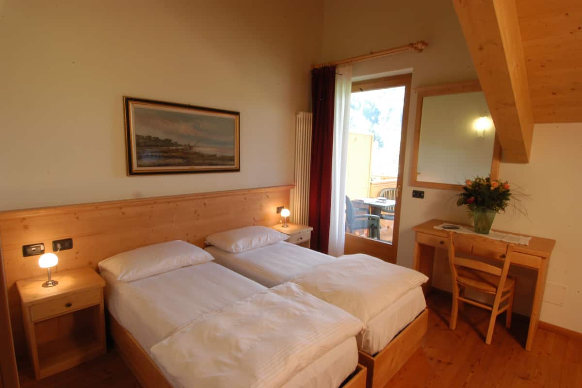 A room with two beds, a desk, a chair, a nightstand, a lamp, a painting, and a window with curtains.