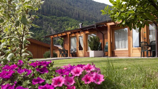 A wooden cabin with a patio area, surrounded by lush greenery and mountains in the background. - Dolomiti Camping Village&Wellness Resort (Dimaro Folgarida)