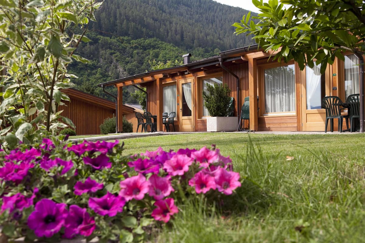 A wooden cabin with a patio area, surrounded by lush greenery and mountains in the background.