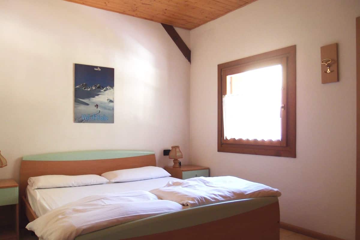A bedroom with a bed, a nightstand, a lamp, a window, and a painting on the wall.