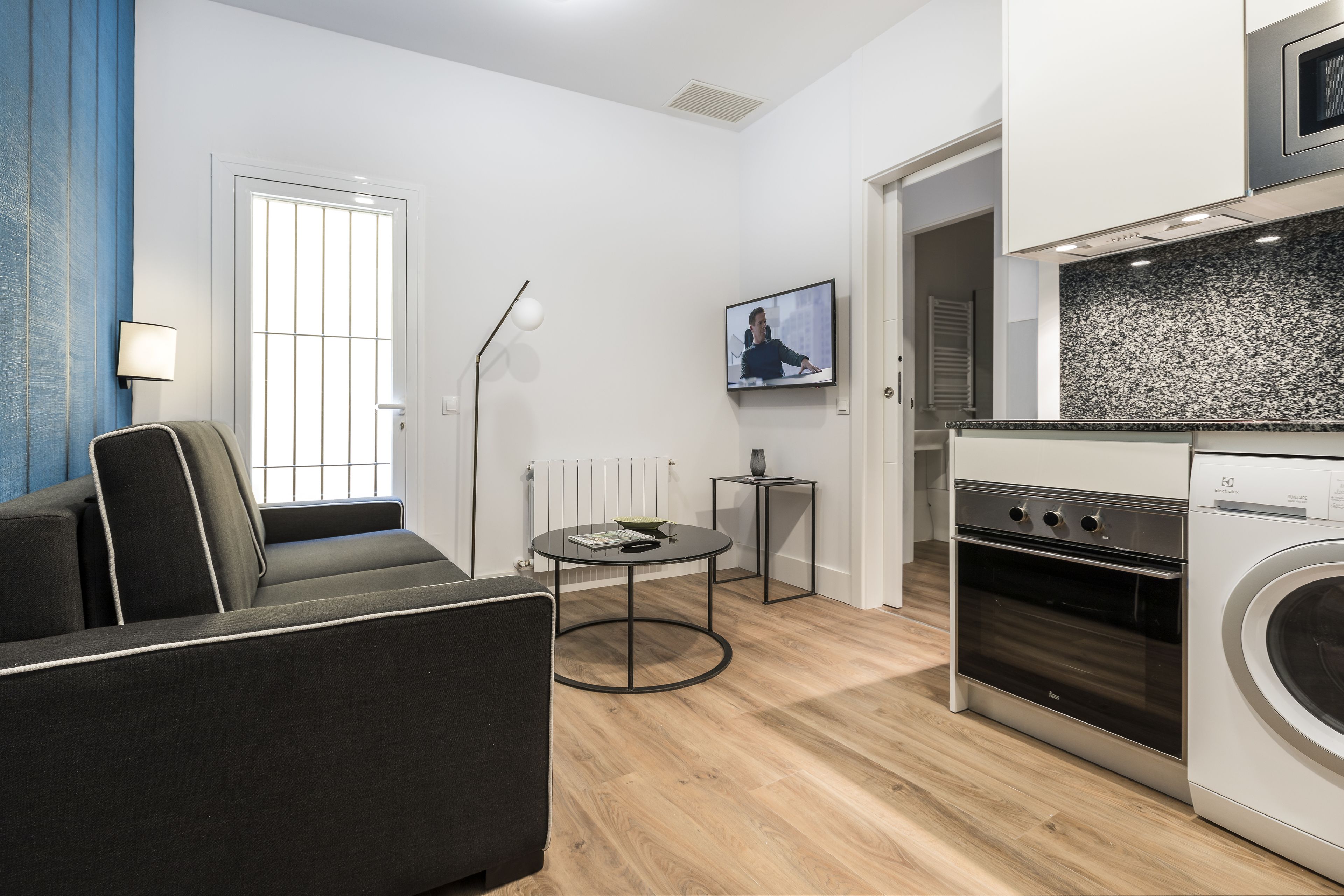 A modern living room with a sofa, a small round table, a TV, and a kitchen area with a washing machine.